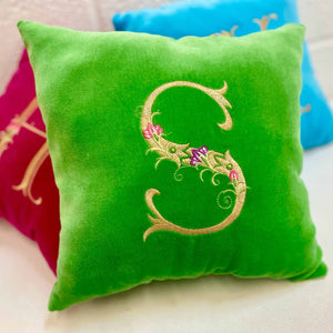 A bright green velvet cushion featuring an ornate gold embroidered monogram with small floral accents, styled on a light surface with other cushions partially visible in the background.