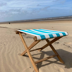 A wooden folding beach chair with a turquoise, white striped fabric seat, set on a sandy beach