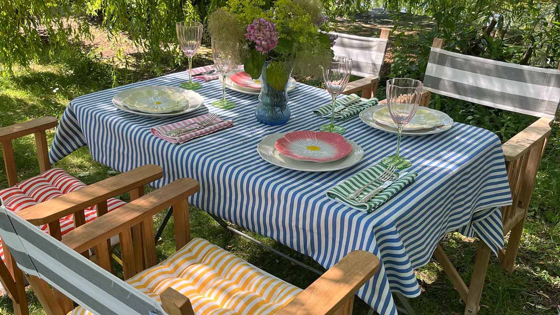Striped tablecloth set on an outdoor dining table with wooden directors chairs, colourful striped napkins and striped seat pads