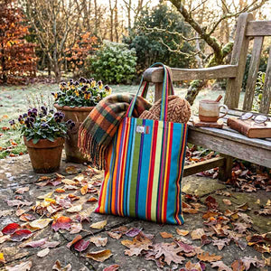 A colourful red, green, blue and yellow vintage print striped cotton tote bag stands on a stone patio outdoors, surrounded by fallen leaves, potted flowers, and a wooden bench