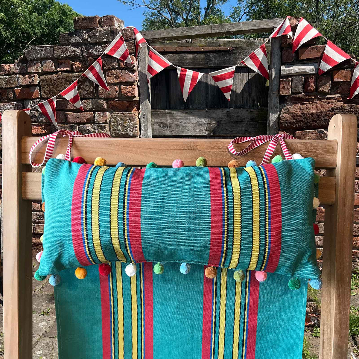 headrest cushion on a wooden chair outdoors, featuring a jade green red and yellow stripe pillow with multicoloured pom-poms and matching striped fabric deckchair sling, with red-and-white bunting