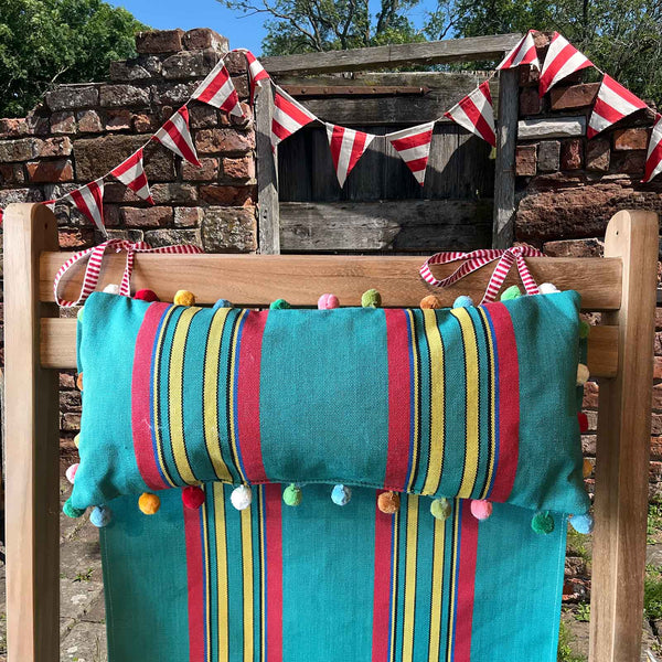 headrest cushion on a wooden chair outdoors, featuring a jade green red and yellow stripe pillow with multicoloured pom-poms and matching striped fabric deckchair sling, with red-and-white bunting