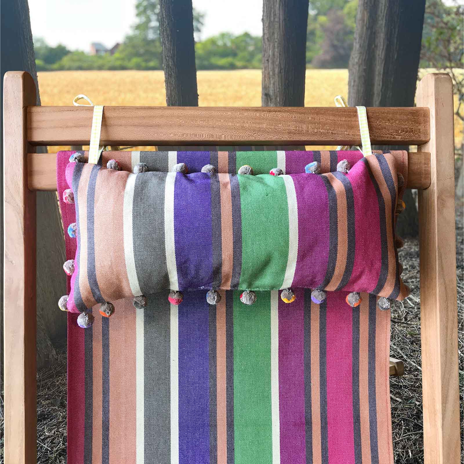 colourful striped headrest lumbar pillow with pom-pom trim hanging from a wooden deckchair