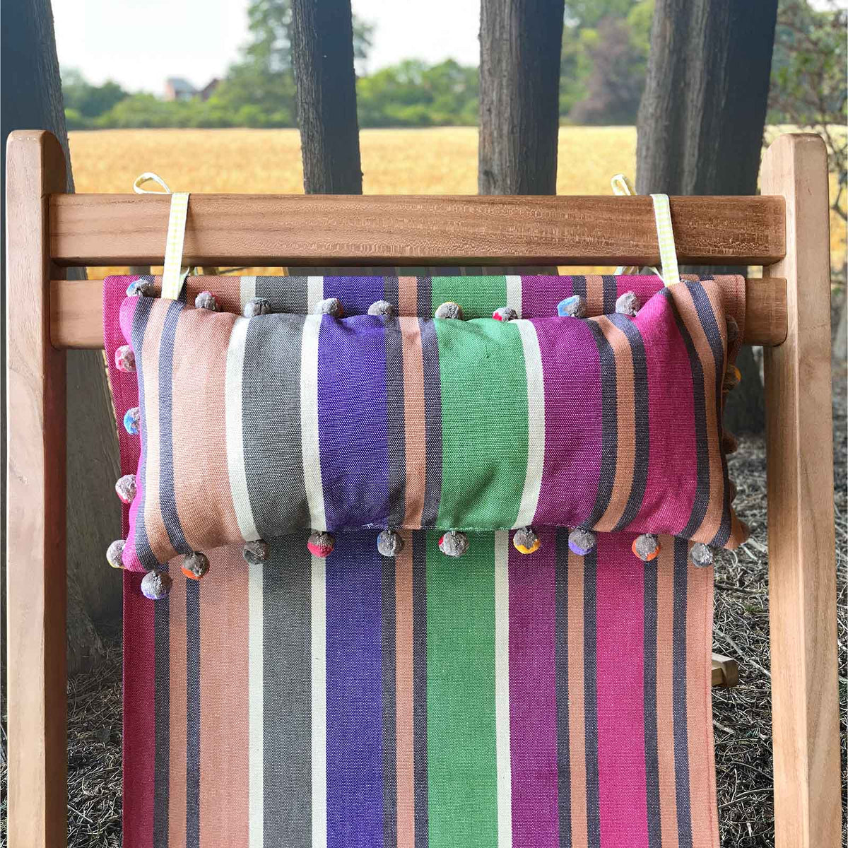colourful striped headrest lumbar pillow with pom-pom trim hanging from a wooden deckchair
