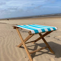 A wooden folding beach chair with a turquoise, white striped fabric seat, set on a sandy beach 