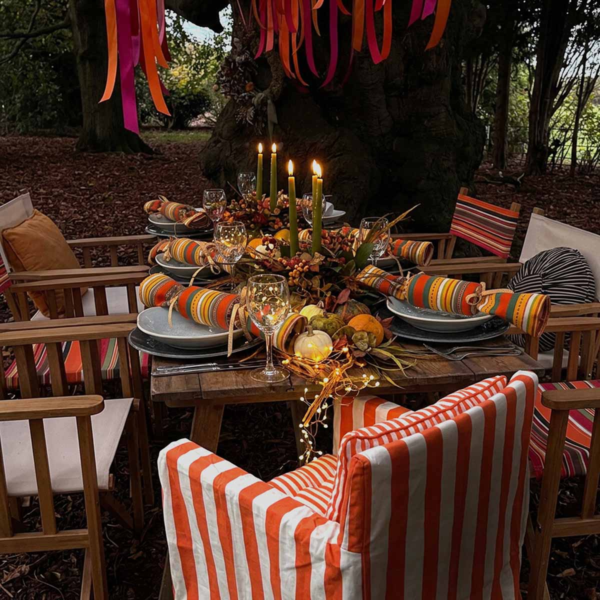 Outdoor autumn table setting under a large tree, featuring striped orange and white chair, colorful napkins, reusable fabric crackers and fall-themed table decor.