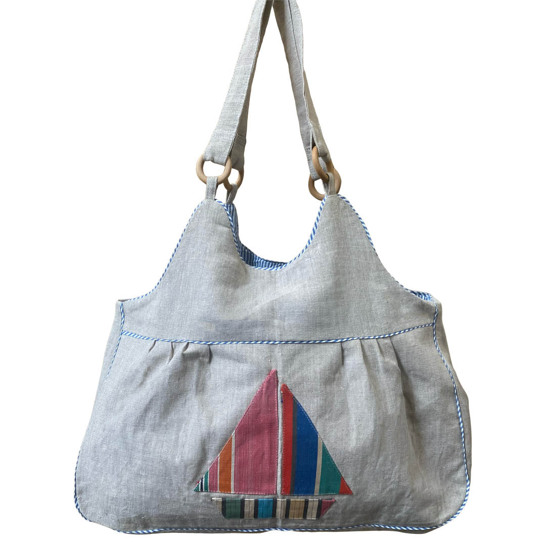 A light gray linen shoulder bag with a colorful striped sailboat applique on the front pocket and blue contrast stitching, shown against a plain white background.