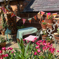 A styled outdoor patio corner with a small table and chair set under a string of colorful striped bunting flags. The table has a green and white striped tablecloth, and the chair has a pink and white striped cushion.