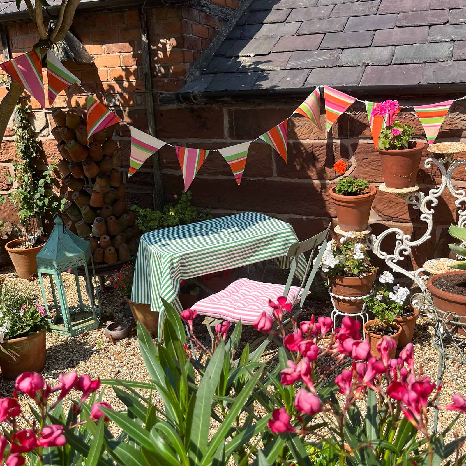 A styled outdoor patio corner with a small table and chair set under a string of colorful striped bunting flags. The table has a green and white striped tablecloth, and the chair has a pink and white striped cushion.