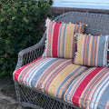 striped cushions in red, beige, grey, and orange, accented with pom-pom trim on a grey wicker outdoor loveseat
