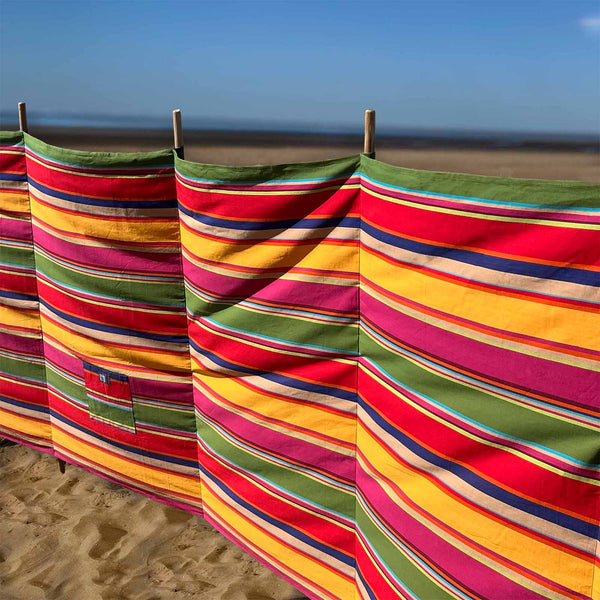 A colourful striped beach windbreak with horizontal bands of red, yellow, green, pink, and blue on a sandy beach with the ocean and blue sky in the background.