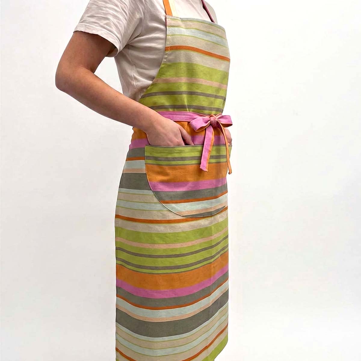 long apron with horizontal pastel stripes in green, orange, pink, beige and grey, tied at the waist with a pink bow. The apron has a large front pocket
