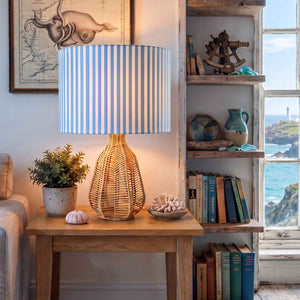 A coastal-style light blue and white striped fabric lampshade on a table lamp in a nautical-themed room with  a window overlooking the sea.