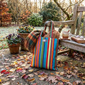 A colourful red, green, blue and yellow vintage print striped cotton tote bag stands on a stone patio outdoors, surrounded by fallen leaves, potted flowers, and a  wooden bench
