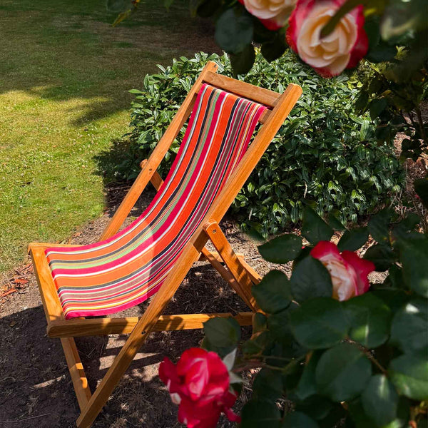 A wooden folding deck chair with a vibrant orange, red, pink, and green striped fabric sling, set in a garden with green foliage and red roses.