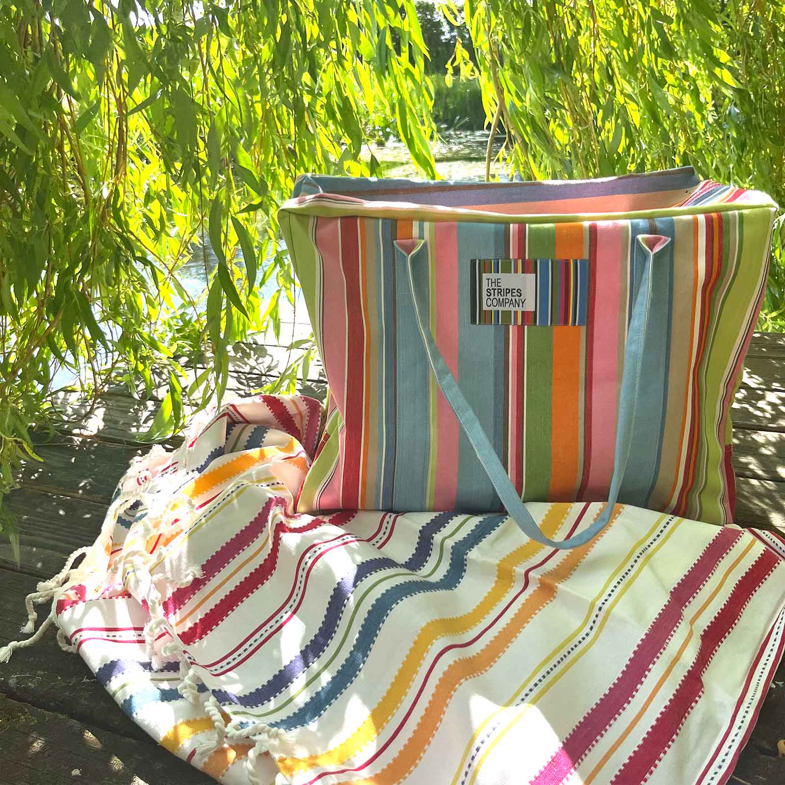 A colorful striped tote bag with light blue handles sits on a wooden bench outdoors under weeping willow branches, next to a matching striped blanket.