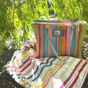 A colorful striped tote bag with light blue handles sits on a wooden bench outdoors under weeping willow branches, next to a matching striped blanket.